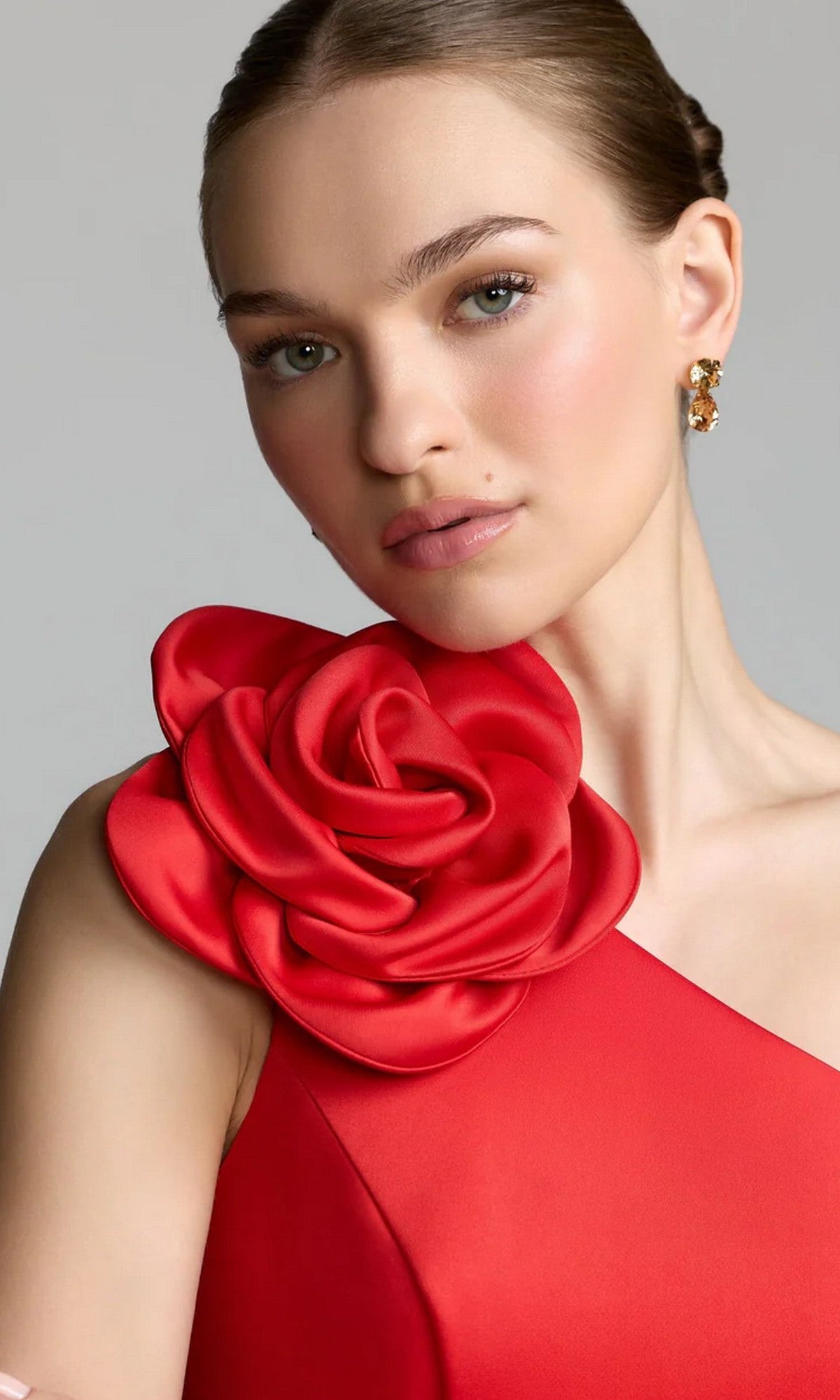 A woman poses against a plain background, wearing the Midi Party Dress: Mac Duggal 2364—a stunning red one-shoulder party dress featuring a large fabric rose on the shoulder—paired with stud earrings.